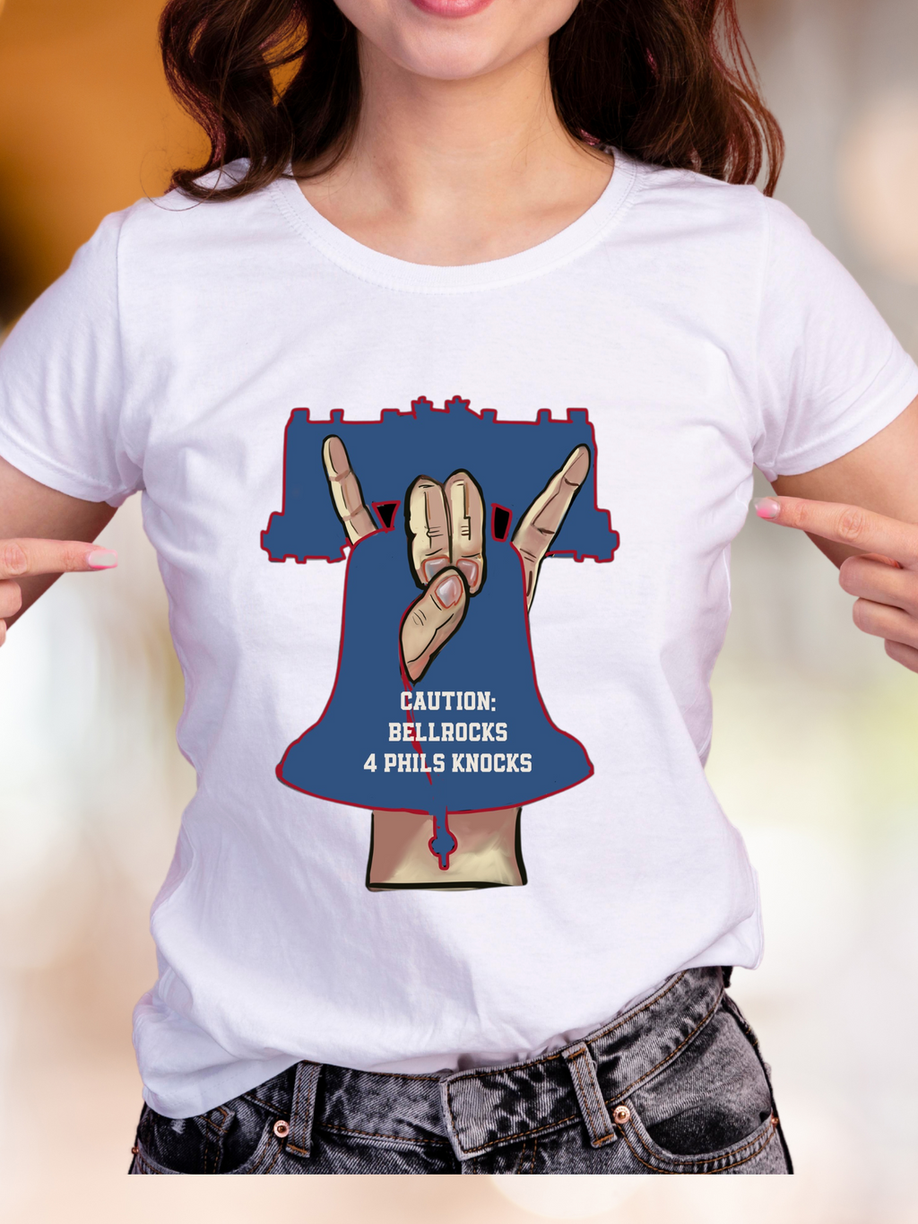 WOMAN IN White t-shirt with a bold graphic of a hand gesture forming a bell shape inside a Liberty Bell outline, with text that reads “Caution: Bellrocks 4 Phils Knocks